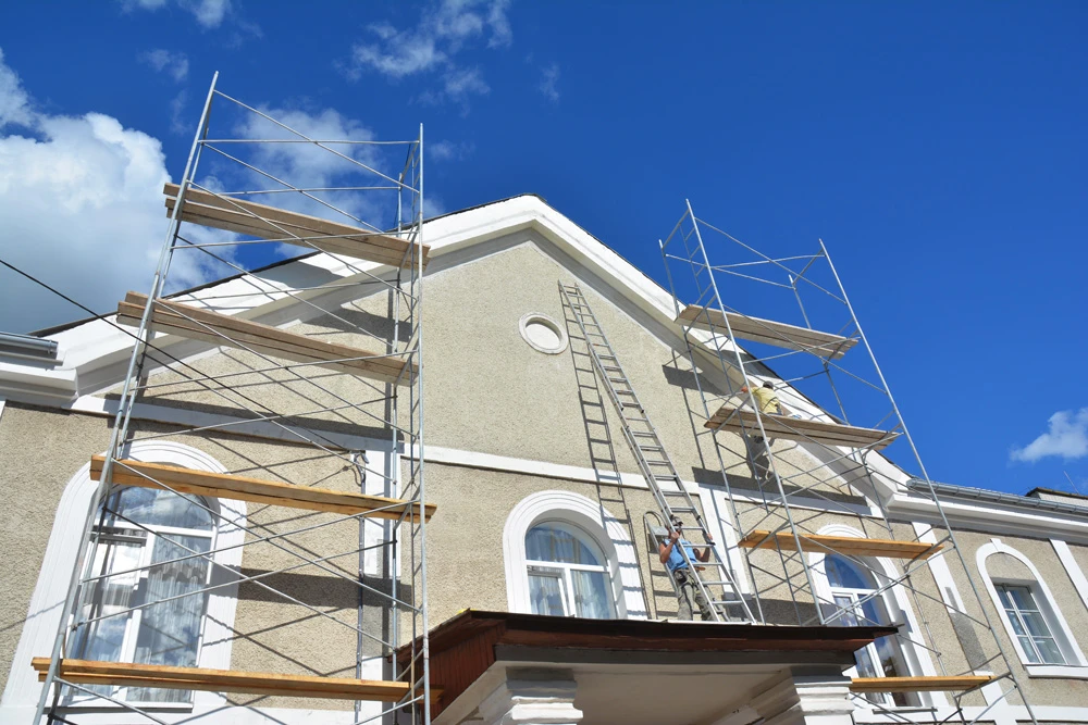 Ouvrier en équipement de sécurité applique une peinture de façade extérieure à l'aide d'un rouleau sur mur blanc d'habitation résidentielle